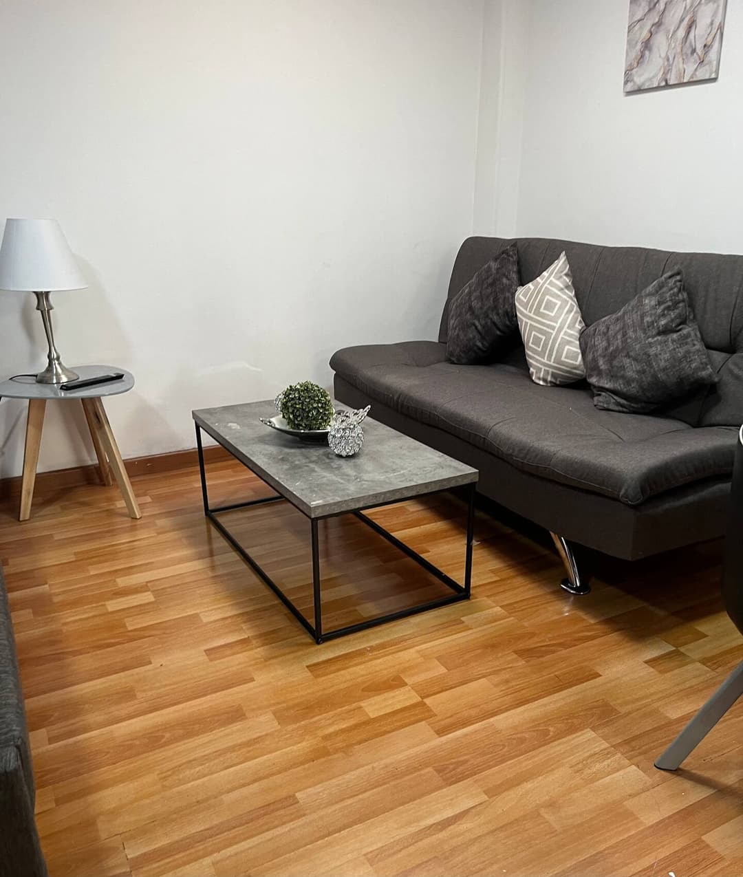 Sala de estar con un sofá gris, cojines decorativos, una mesa de centro de diseño minimalista y una lámpara de mesa blanca sobre un pequeño mueble auxiliar. El suelo es de madera.