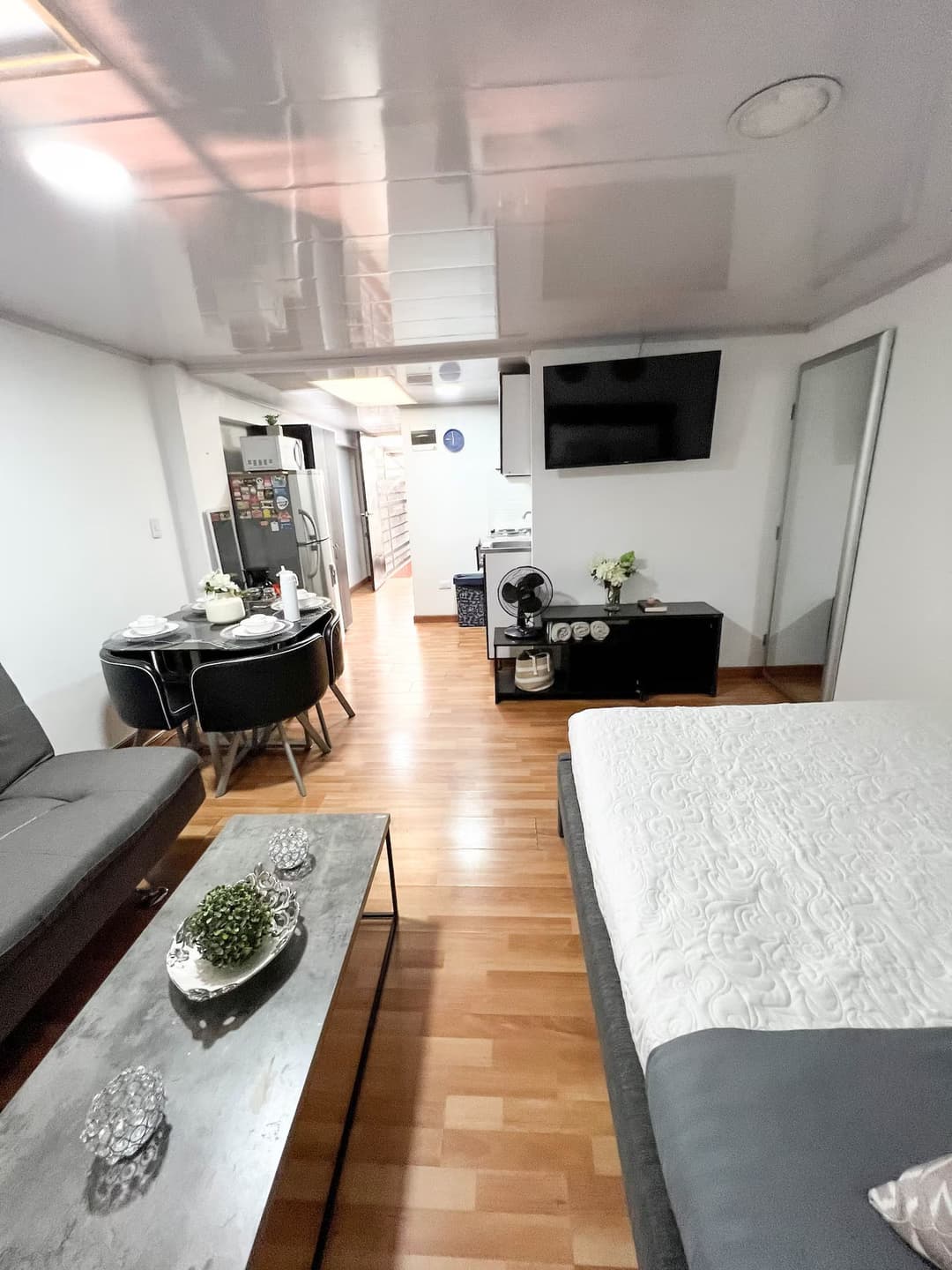 Vista de un espacio abierto que combina sala de estar y cocina. Se observa un sofá, una mesa de centro con decoración, una cama, una televisión montada en la pared y una mesa de comedor con sillas negras.