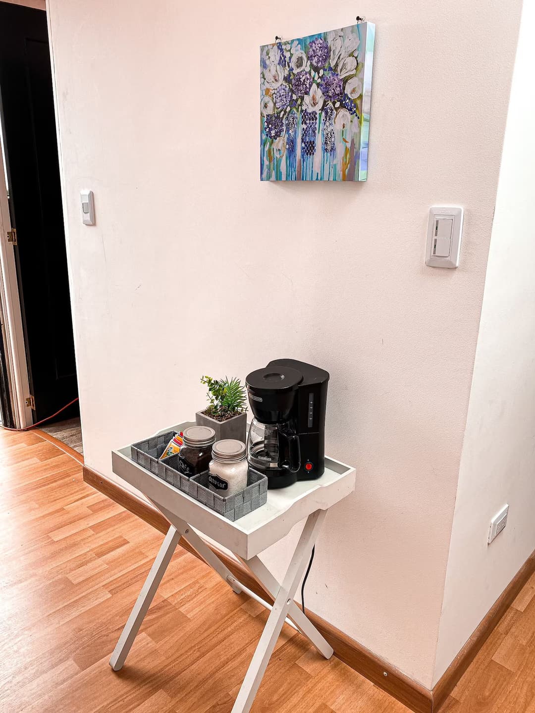 Esquina de una habitación con una cafetera sobre una mesa blanca plegable. En la pared hay un cuadro colorido con flores.