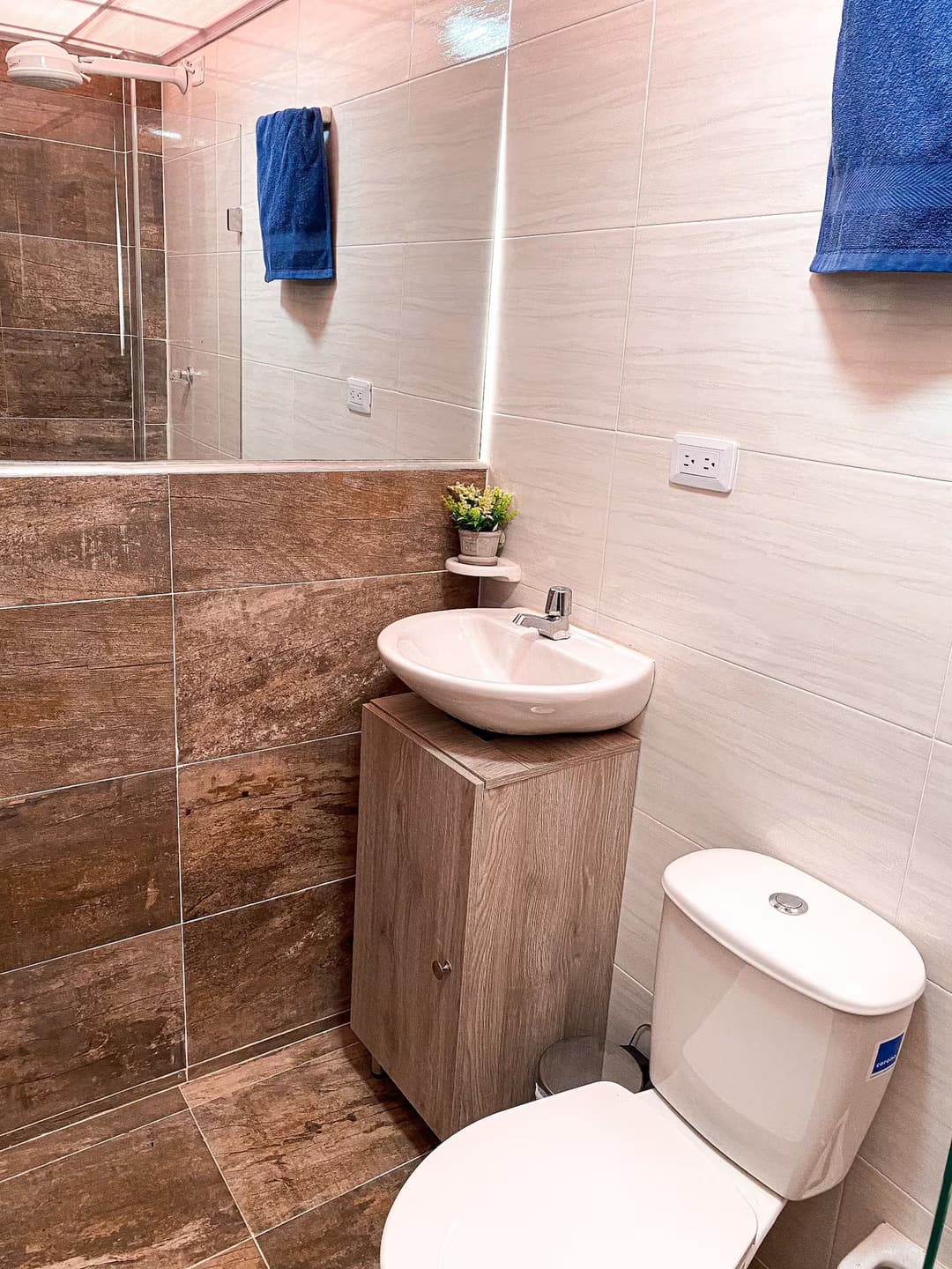 Baño moderno con azulejos marrones y blancos. Incluye un lavabo sobre un mueble de madera, un inodoro y una toalla azul colgada en la pared.