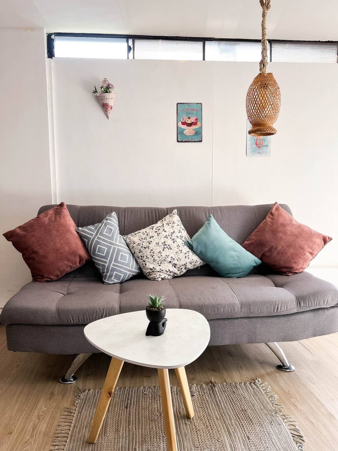 Una sala moderna con un sofá gris adornado con cojines multicolores. Una mesa de centro blanca con patas de madera sostiene una pequeña planta decorativa. Las paredes tienen decoración minimalista y hay una lámpara colgante de mimbre.