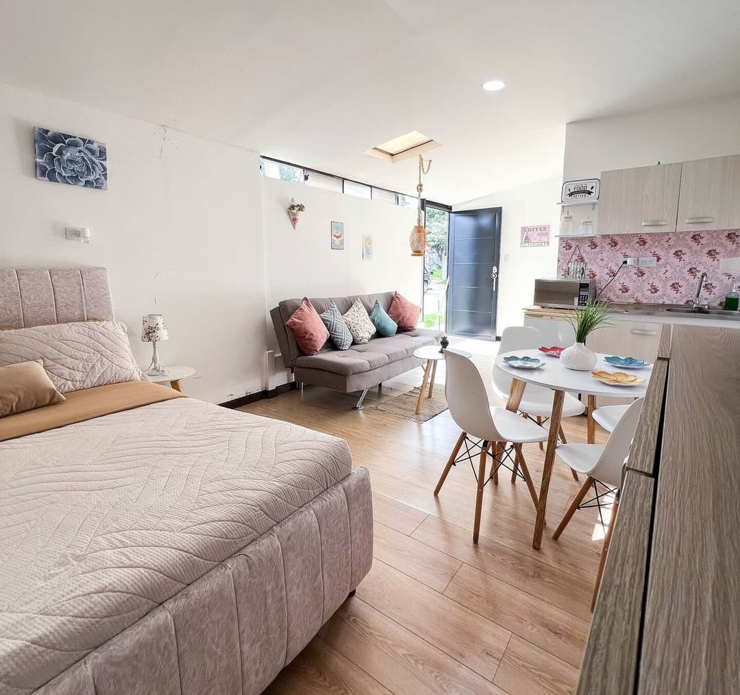 Un espacio abierto que combina una sala de estar, cocina y dormitorio. Cuenta con un sofá gris, una cama cómoda y un comedor con sillas blancas. La cocina es compacta y tiene un fondo floral. Un ambiente acogedor y luminoso.