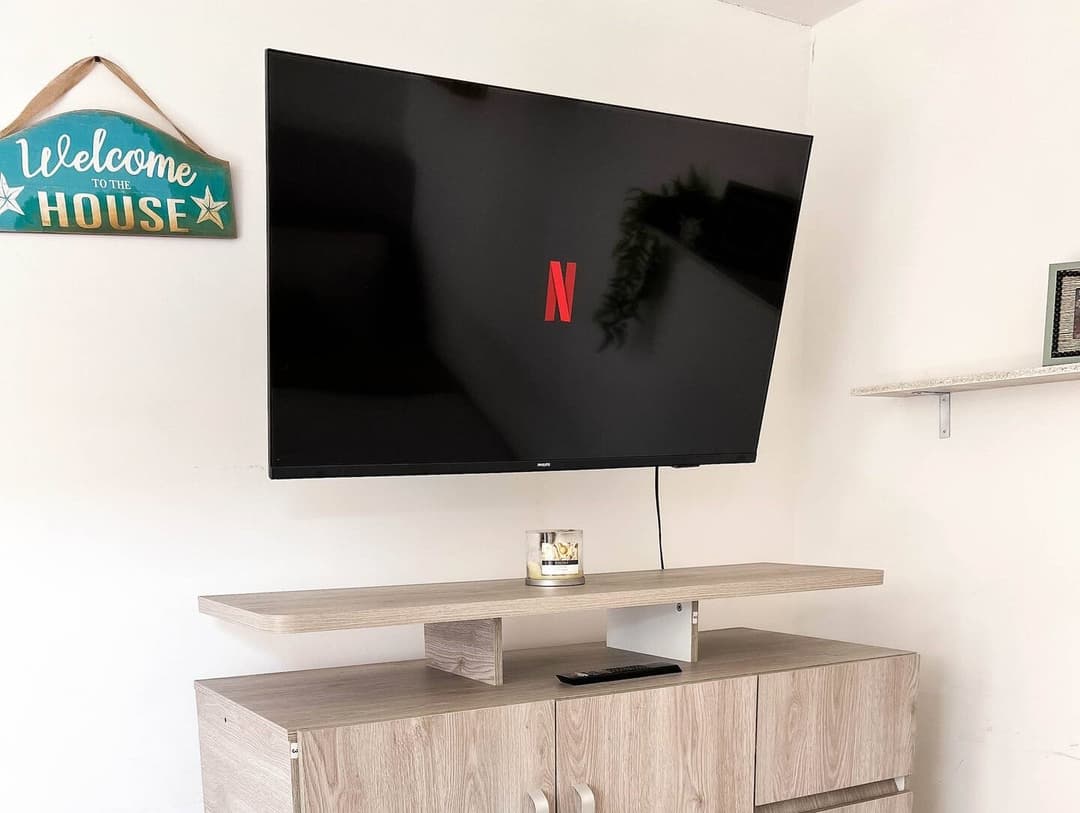 Un televisor montado en la pared sobre un mueble de madera clara. A su lado, un cartel de bienvenida en la pared. El ambiente es moderno y minimalista.