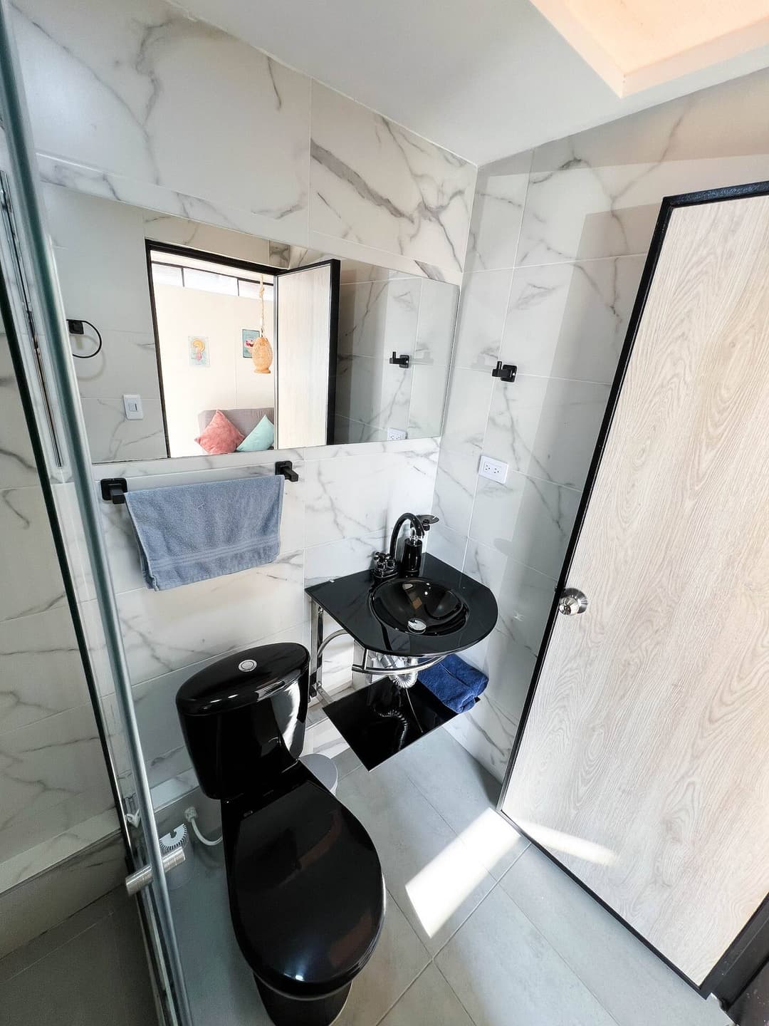 Un baño elegante con azulejos de mármol claro, equipado con un mueble de lavabo negro y un inodoro negro. La iluminación natural entra por una ventana pequeña, proporcionando un ambiente luminoso y moderno.