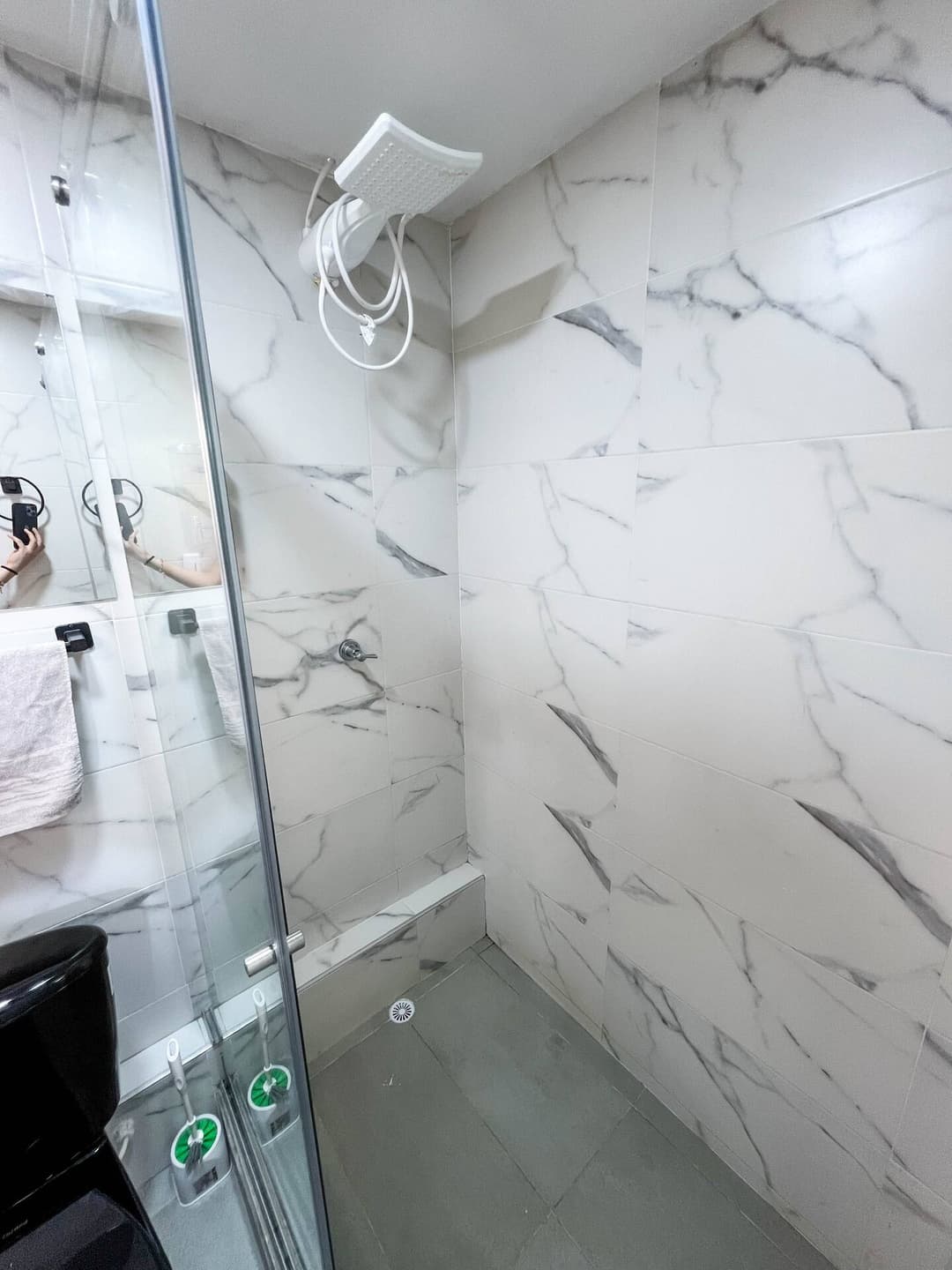 Un cuarto de baño con azulejos de mármol y una ducha moderna con cabezal blanco. La mampara de vidrio muestra el reflejo de las toallas y otros accesorios.
