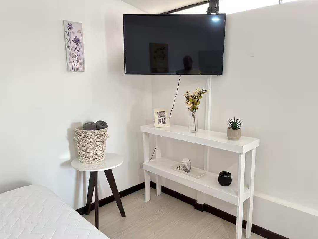Sala Acogedora: Un televisor montado en la pared sobre una mesa blanca estrecha con decoración mínima. A su lado, una mesa redonda con un cesto decorativo y una decoración de pared con flores.