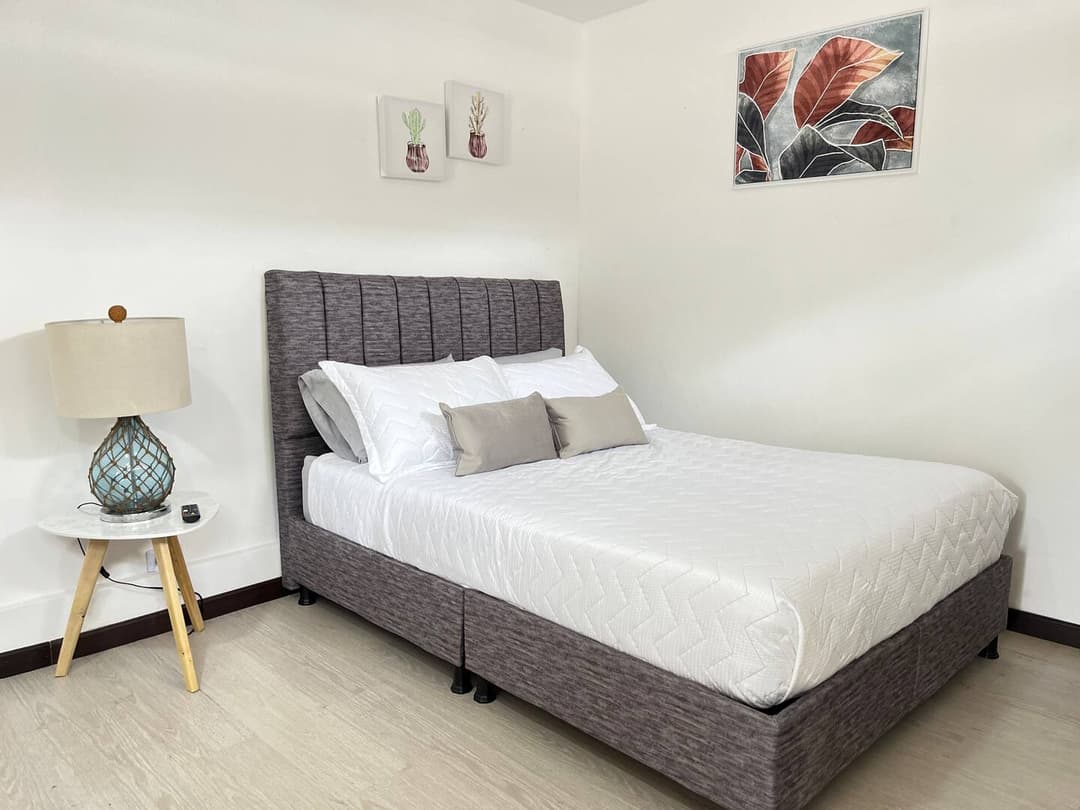 Dormitorio Elegante: Una cama de diseño moderno con un cabecero acolchado gris, cojines y ropa de cama blancos. Una mesita de noche con una lámpara y cuadros decorativos que adornan la pared.