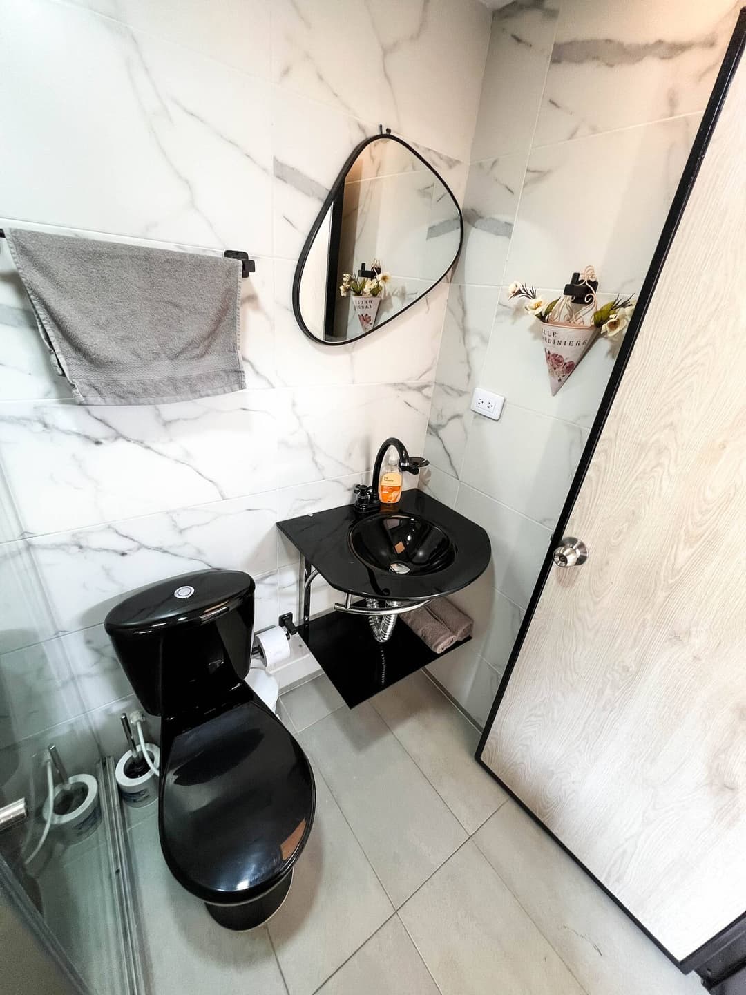 Baño Moderno: Un baño pequeño y elegante con azulejos de mármol, inodoro y lavabo negros. Un espejo de forma ovalada cuelga sobre el lavabo, y una toalla gris está doblada en un riel junto a la pared.