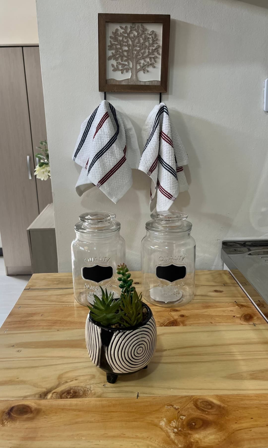 Detalle de Cocina: Encimera de madera con frascos de vidrio y pequeñas plantas decorativas. Dos paños de cocina cuelgan de un perchero, con un cuadro de un árbol sobre ellos, añadiendo un toque cálido.
