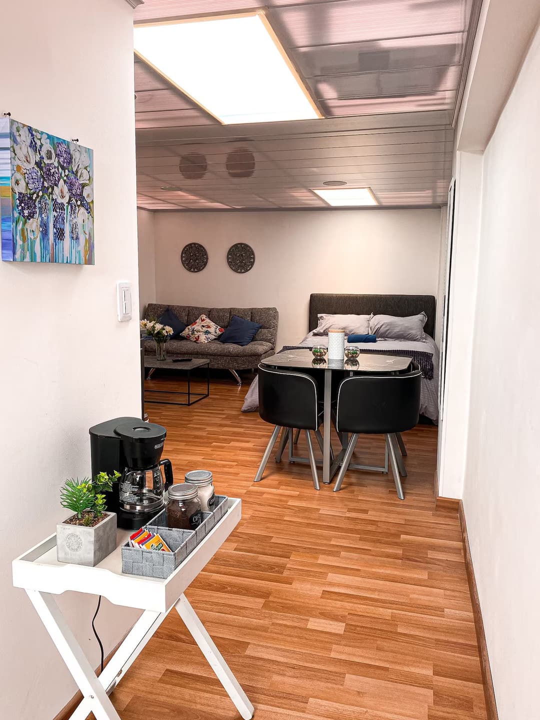 Entrada a un área común con una cafetera sobre una pequeña mesa blanca. Al fondo, una sala de estar y un área de comedor con sillas negras y un sofá decorado con cojines coloridos.
